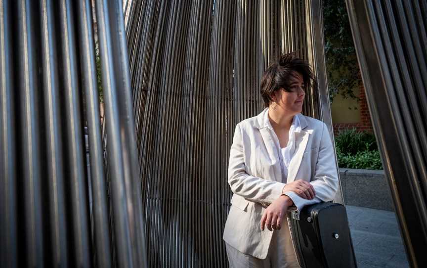 A person in a cream casual suit stands in front of an abstract building leaning on a guitar case